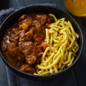 Rindergulasch mit Spätzle, serviert in einer Schüssel auf dunklem Hintergrund. Nahaufnahme.